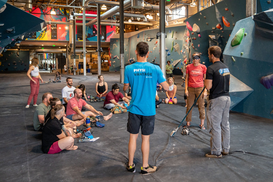 Set inside a bouldering gym, backs facing cameras - Justin Salas addresses a group of individuals attending a climbing clinic - sat on the floor in front of him. To his right, Nate and Seneida (blindfolded) are demonstrating the fundamentals of communications between a climber and their caller.
