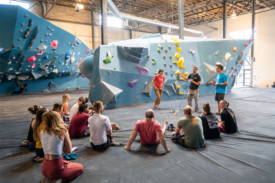 Backs facing camera %u2013 a group of people sit on the floor of a bouldering gym facing Justin Salas, Nate, and Seneida (blindfolded). Nate is explaining the fundamentals of communications between a caller and a low vision or blind climber.