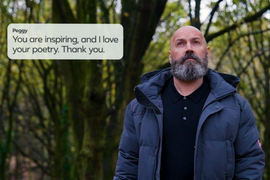 Taken from Dave's latest work for Meta – shot from waist up, Dave looks upwards towards the sky (head on to camera). His expression is one of deep thought and reflection. He wears a navy blue down jacket with a black button up polo beneath. Behind in and out of focus, green foliage of a forest. Top left of frame, a speech bubble reads a comment by one of Dave's fans, Peggy: You are inspiring, and I love your poetry. Thank You.