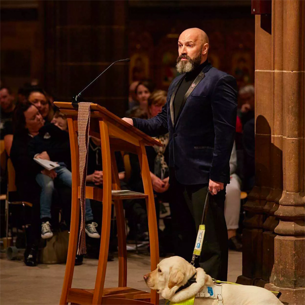 Dave stands next to a wooden lectern, presenting in a blue velvet tuxedo. His guide dog stands by his side.