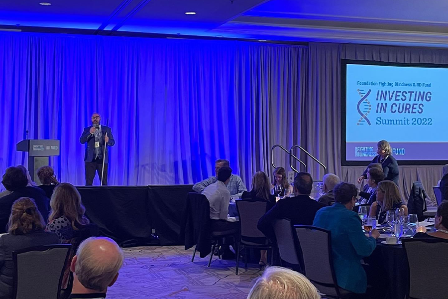 Shot from afar – Dave stands on stage speaking at the Investing in Cures Summit 2022; held by the Foundation Fighting Blindness & RD Fund. Backs turned to the camera in the midground and foreground, a room full of guests are seated around tables adorned by wine glasses and plates. A blue spotlight floods the stage where Dave stands.