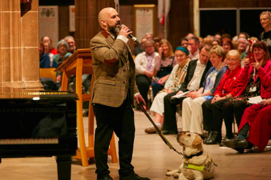 Dave speaking at an event in a large hall filled with people; many smiling and looking in Dave's direction. His guide dog sits on the floor and looks up at him. Behind him, out of focus and half in frame, a black grand piano.