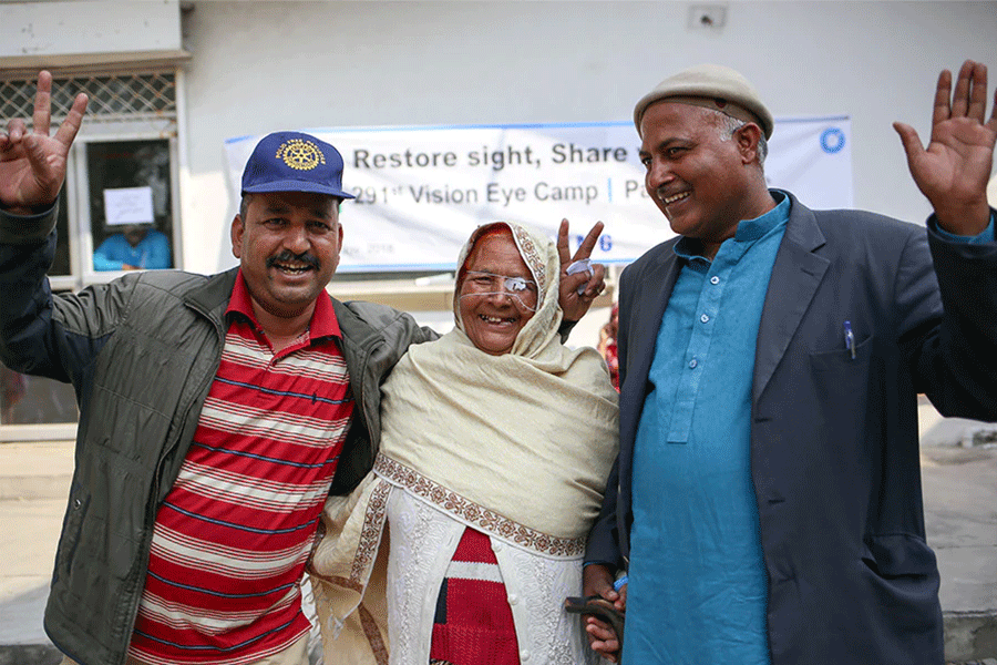 This scene was taken from Vision Care US' eye camp hospitals, offering free eye surgery, check ups, and training for local doctors in various locations around the world. Three individuals; two middle aged gentlemen and an elderly lady between them, look happily at the camera. The gentleman on the left wears a horizontally striped red shirt, under a muddy khaki green jacket and a blue Lions Club International baseball cap. The lady; centre, wears traditional Afghan wear, in cream white and red. She has a transparent eye patch across her left eye from a recent surgery. The gentleman on the right wears a light blue traditional Afghan shirt and grey jacket.