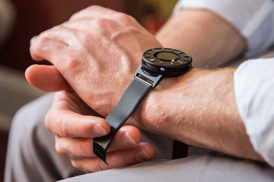  Shot close up: The Eone Bradley Black Mesh rests on a man's left wrist. Around the edge of the image we can see he is wearing a white dress shirt, sleeves rolled, and gray suit pants.