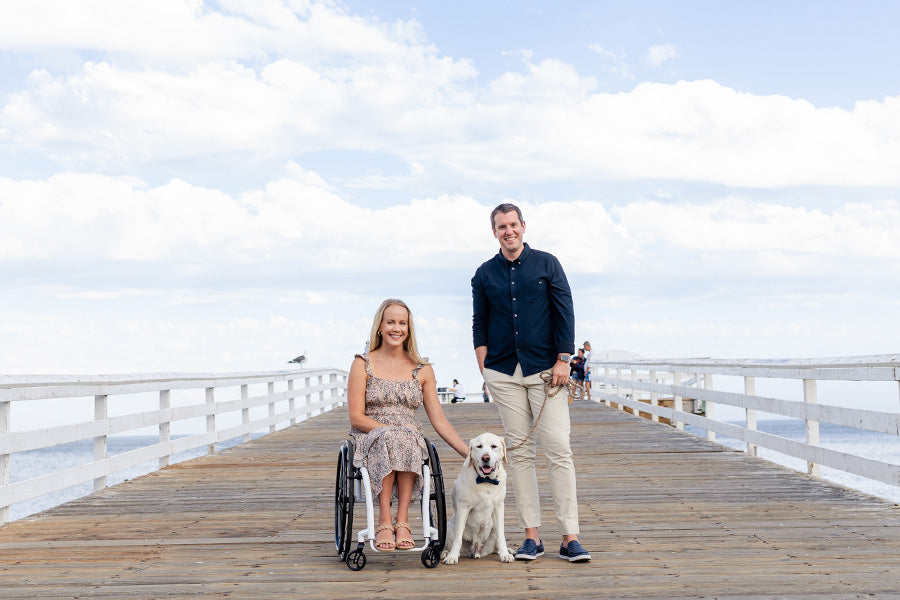 Jeremy, Mallory, and Sam pose and smile at the camera, on a boardwalk. Jeremy wears a navy blue shirt, cream chinos, and leather boat shoes. Mallory wears a speckle floral dress, and sand colored sandals with woven plait details.