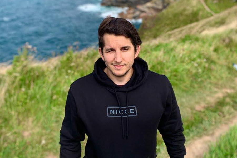 Oliver smiles at the camera stands against green grass hills along a coast wearing a black drawstring hoodie with a white logo along the chest. It appears that he might be winking but it could just be windy!
