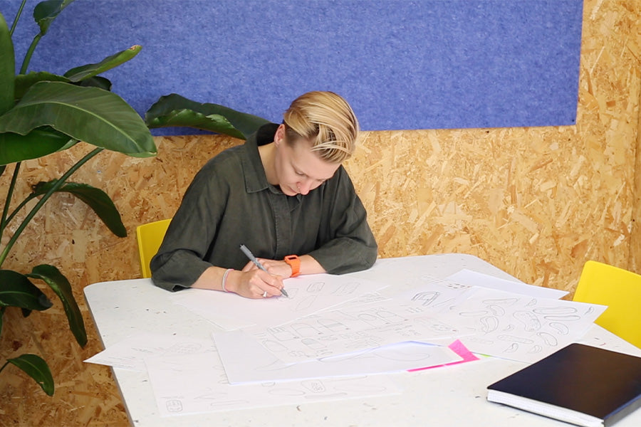 Morrama’s creative director, and founder, Jo Barnard sketches designs on a large white table, seated in a bright yellow chair. Behind her, a chipboard wall with a lilac coloured, felt pinboard. Jo wears a dark leaf green shirt and orange watch.