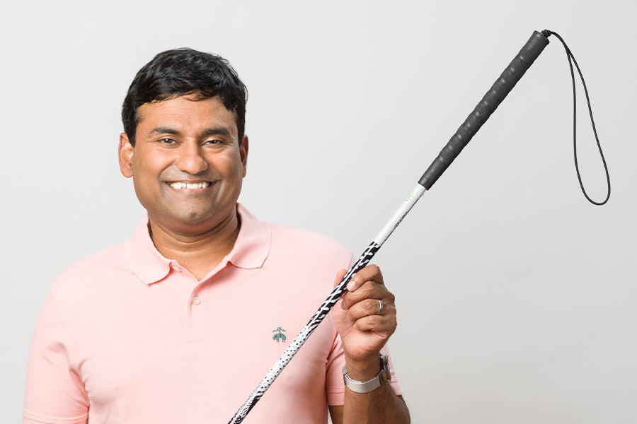 John Samuel, smiling brightly in a light pink polo, showing off his white cane that is wrapped in a unique design