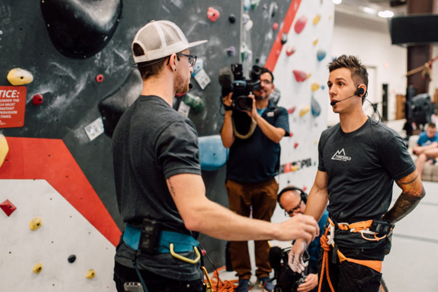 Justin and his climbing partner Matt talks strategy inside a climbing gym, surrounded by spectators. Both are wearing black t-shirts and a cameraman can be seen in the background between them.