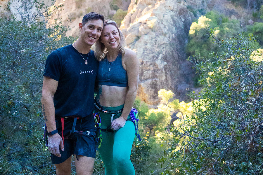 Justin and his girlfriend April, smile at the camera, green foliage and a rocky mountain can be seen in the background. Justin wears a black t-shirt and shorts, April wears a blue sports top and green sports pants.