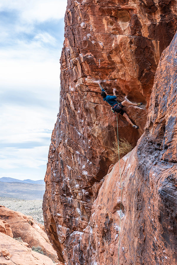 Taken from below looking up - Justin scales a red rock face, wearing a blue long-sleeve top and black climbing shorts. A bright green climbing rope can be seen connected to his harness.