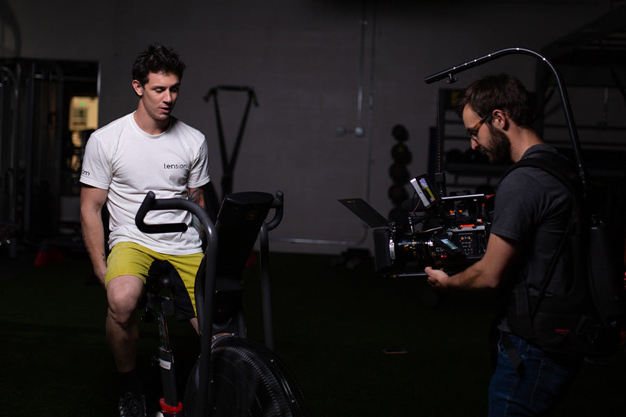 Justin trains on a spinning bike, wearing a white t-shirt and yellow shorts.  A camera man is seen on the right side, recording him.