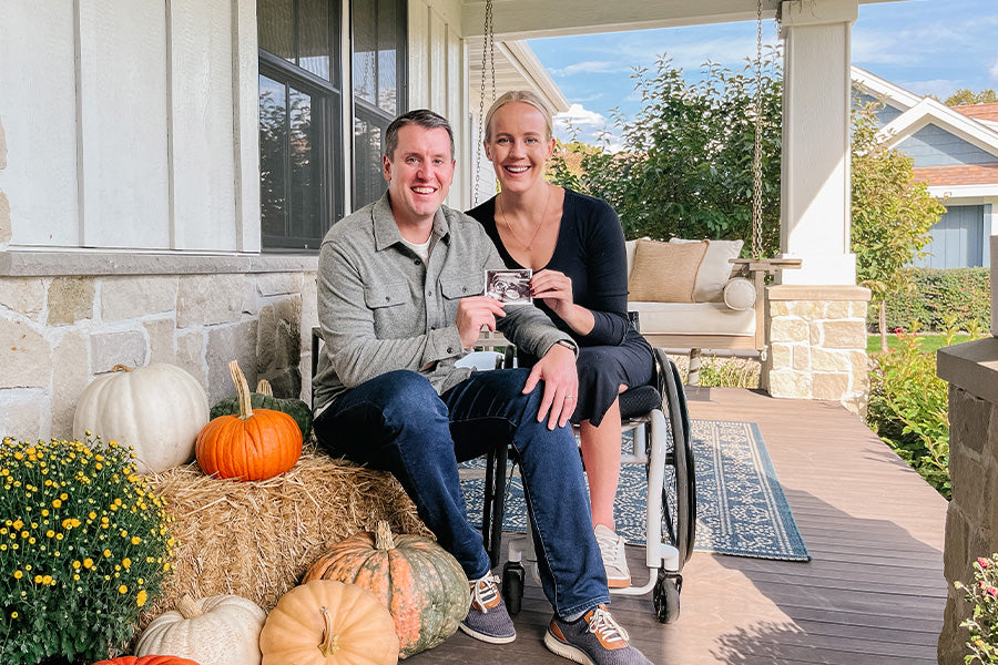 Jeremy and Mallory sit outside their house, smiling ecstatically at the camera. Together, they hold an ultrasound from their successful IVF journey. To their left, pumpkins of various sizes and colors can be seen.