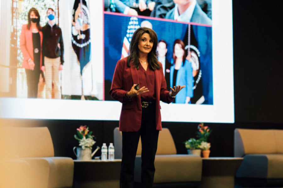 Sharon on stage on her Hearts and Minds talk. She wears a burgundy jacket and blouse, with black pants. Behind her and out of focus, a projector screen of images showing Sharon with different individuals.