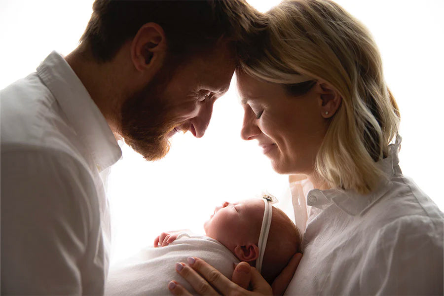 Photoshoot against a white background. Brad and Sara lean into each other, smiling. Between them, held by Brad, a sleeping Baby Rooney wrapped in a white blanket. 