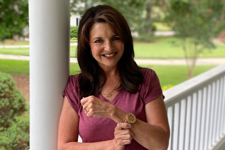 Smiling at the camera, Sharon shows off her new Eone Switch in Rose Gold. She leans against a white support beam with white fencing and green foliage behind her. Sharon wears a pink t-shirt.