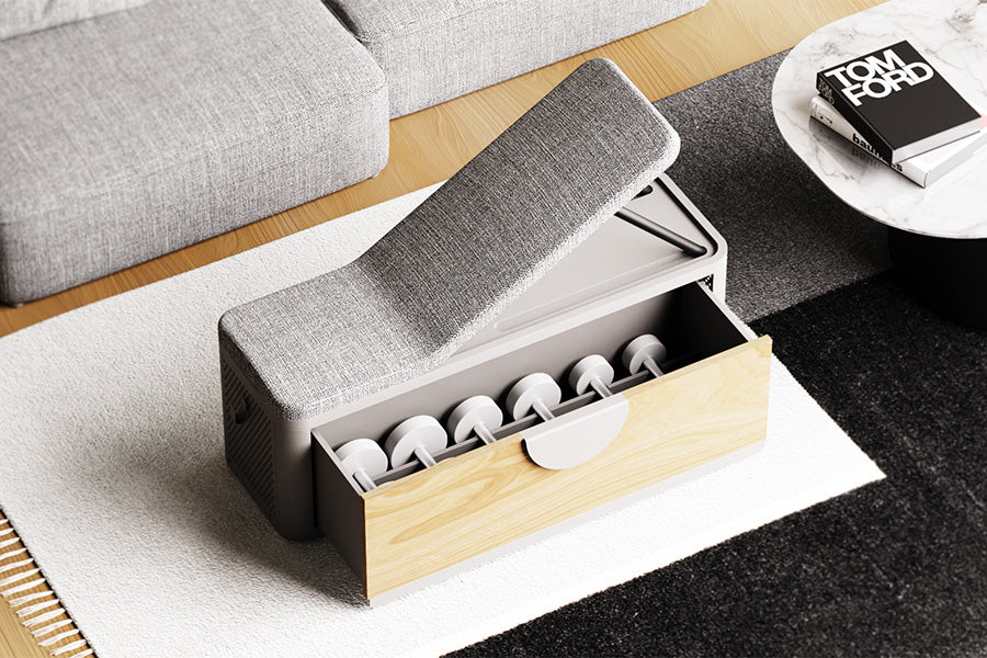 From this perspective, the durable, grey upholstered backrest of the concept exercise bench is set to an incline position. The storage drawer is open towards the viewer, highlighting the light wood front panel and large semi-circular, folded steel handle. The base of the exercise bench is a light, warm stone grey. Oliver’s bench sits atop a white, grey, and black rug, the workout bench upholstery matches that of the sofa in the background. To the top right, a low level coffee table made of marble with a stack of books placed on top.