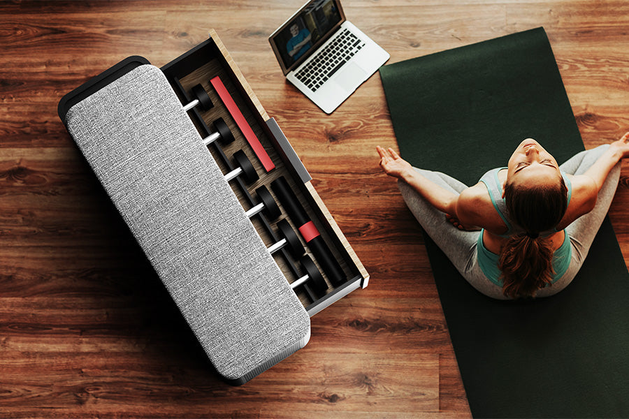 Shot from above, Oliver’s concept exercise bench against a rich brown hardwood floor, with the storage drawer open; revealing a set of dumbbells and accessories. To the right, a lady sits in the lotus position on a yoga mat, her laptop facing her at the edge of the mat. 