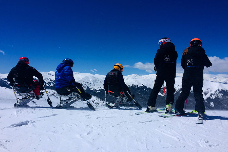 Five athletes from the National Sports Center for the Disabled (NSCD) Competition Team backs faced to the camera, in ski and snow board gear. In the background, white snow capped mountains and a rich blue sky.