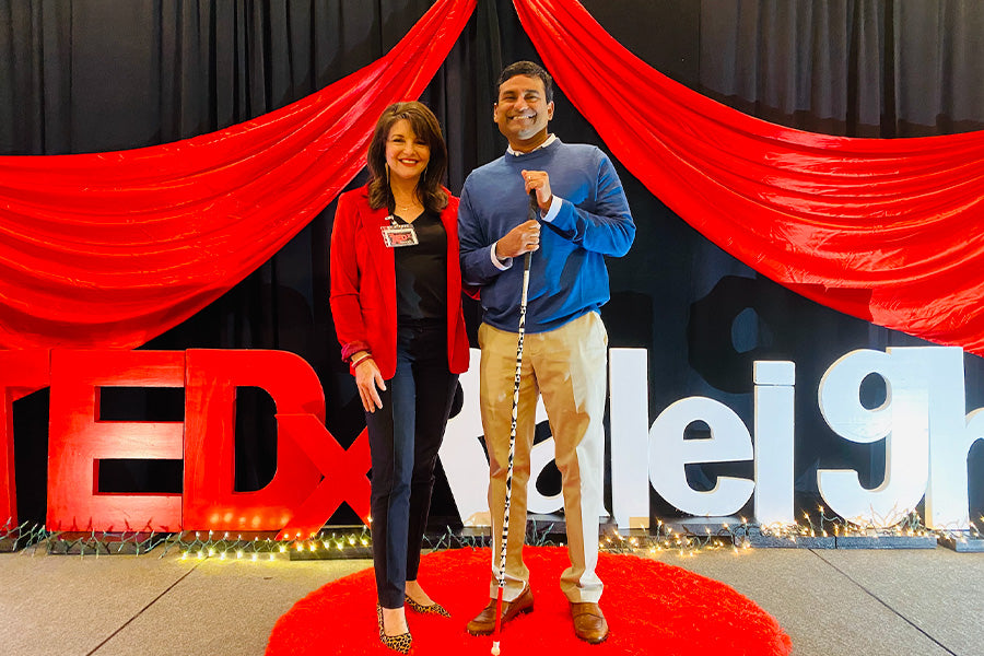 Sharon and John Samuel smile at the camera, standing on stage at Tedx Raleigh. Sharon wears a red jacket, black t-shirt, and black dress pants. John wears a white dress shirt under a blue crew neck knit, beige suit pants and tan brown loafers. John holds the winning design from last year's Drip My Cane project. Click the image to read about our joint efforts for White Cane Day 2021.