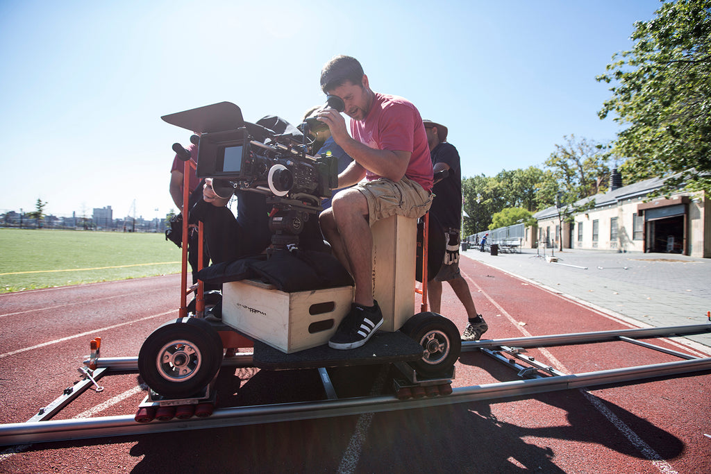 Film-maker Chris Suchorsky sits on a dolly mounted camera rig, looking into the viewfinder. The crew are setup on a running track, blue skies and sunshine backlight the individuals.