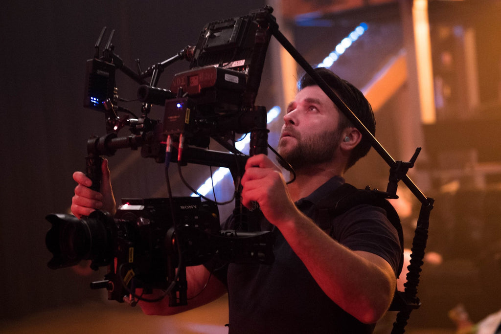 Film-maker Chris Suchorsky holds a camera rig up high, his face lit by the reference monitor. The whole scene is cast in a red and yellow light.