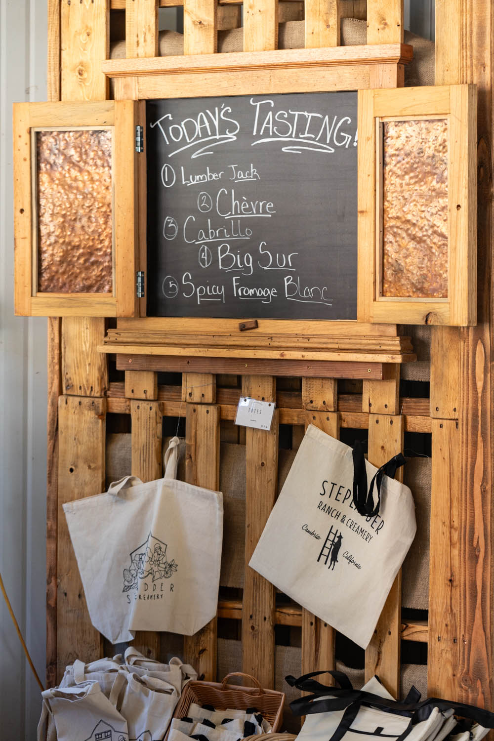 Chalkboard with cheese tasting menu and reusable creamery tote bags displayed below.