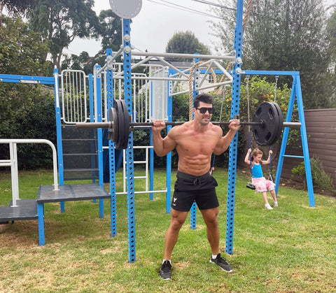 Fit father does back squats on outdoor home gym with daughter swinging nearby