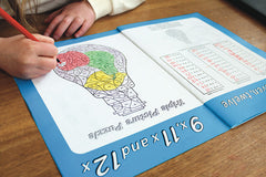 A book is open on the table and a child is facing towards us colouring in. The left side of the book has a light bulb shaped picture divided into sections with a number on each. The right side of the page has a series of sums in the 9,11 and 12 times tables.