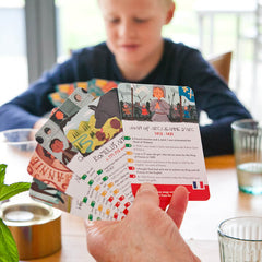 A close up of a hand of History Heroes: Children cards is in the foreground. Joan of Arc is the top card, showing a hand drawn picture of her and 7 facts listed underneath. These are divided into red, amber and green facts for difficulty, and one 'Joker Fact', A child is in the background holding a hand of cards.