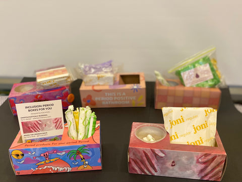 Photo of 5 period boxes, two in a front row and three in a back row. There are pads, tampons and postcards in the boxes.