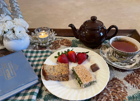 The top of a tea table with a tablecloth, cucumber sandwich, strawberries, and chocolate on a plate, black tea in a teacup, a Brown Betty teapot, a candle, a book, and other decor.