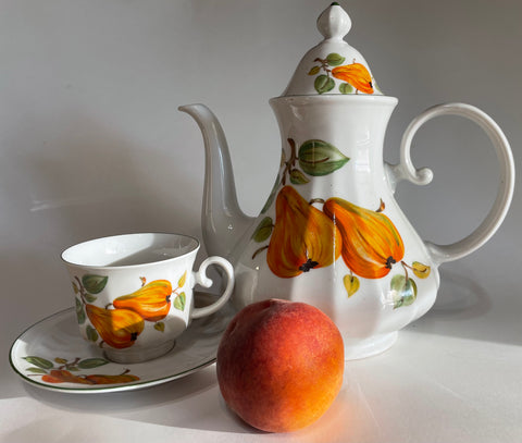 A ceramic white teapot and adjacent cup and saucer painted with an orange fruit design. A peach is in front of the teapot.