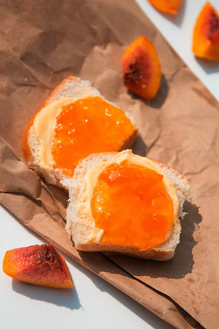 Two slices of bread spread with butter and peach jam on top of a brown paper bag.