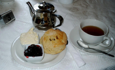 A raisin scone provided with red jam and clotted cream. A silver teapot and white ceramic cup and saucer with black tea are behind the scone.