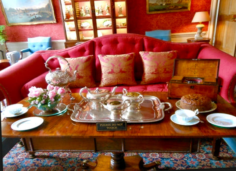 An elegant living room with a red couch and tea table. A silver tea set is on the table along with a fruit cake.