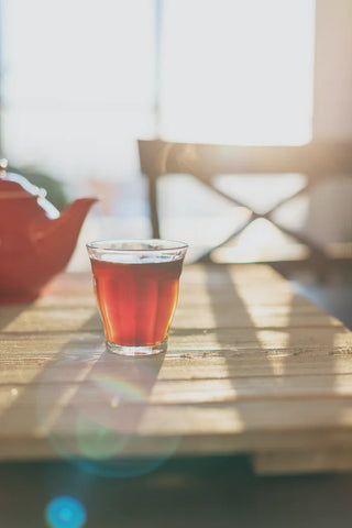 A glass of iced tea on a wooden table, with a red ceramic tea pot in the background.
