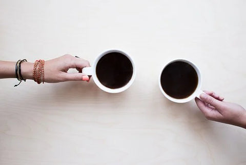 Two people, each holding a cup of strong black tea set against a white table. The hand and wrist of each person is shown.