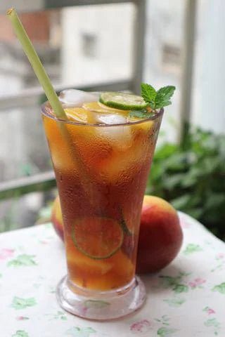 A glass of iced tea on a table next to some apples. The iced tea has a straw, lime slices, and pineapple pieces.