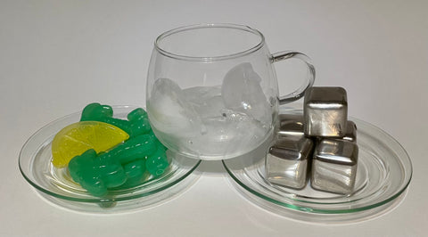 A glass teacup with ice cubes balancing on two saucers. One saucer has plastic palm trees and a plastic lemon slice on it, and the other has stainless steel cubes.