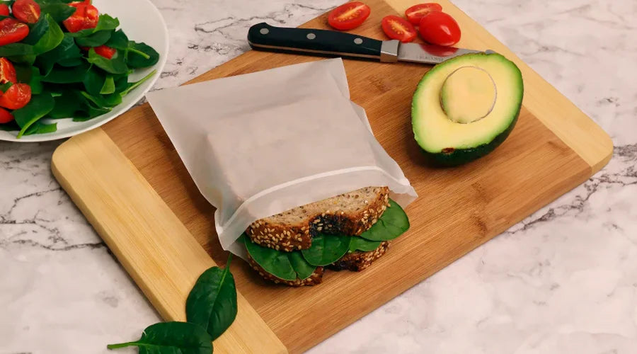 A pla ziplock bag accompanies a sandwich, knife, and a bag of lettuce on a cutting board