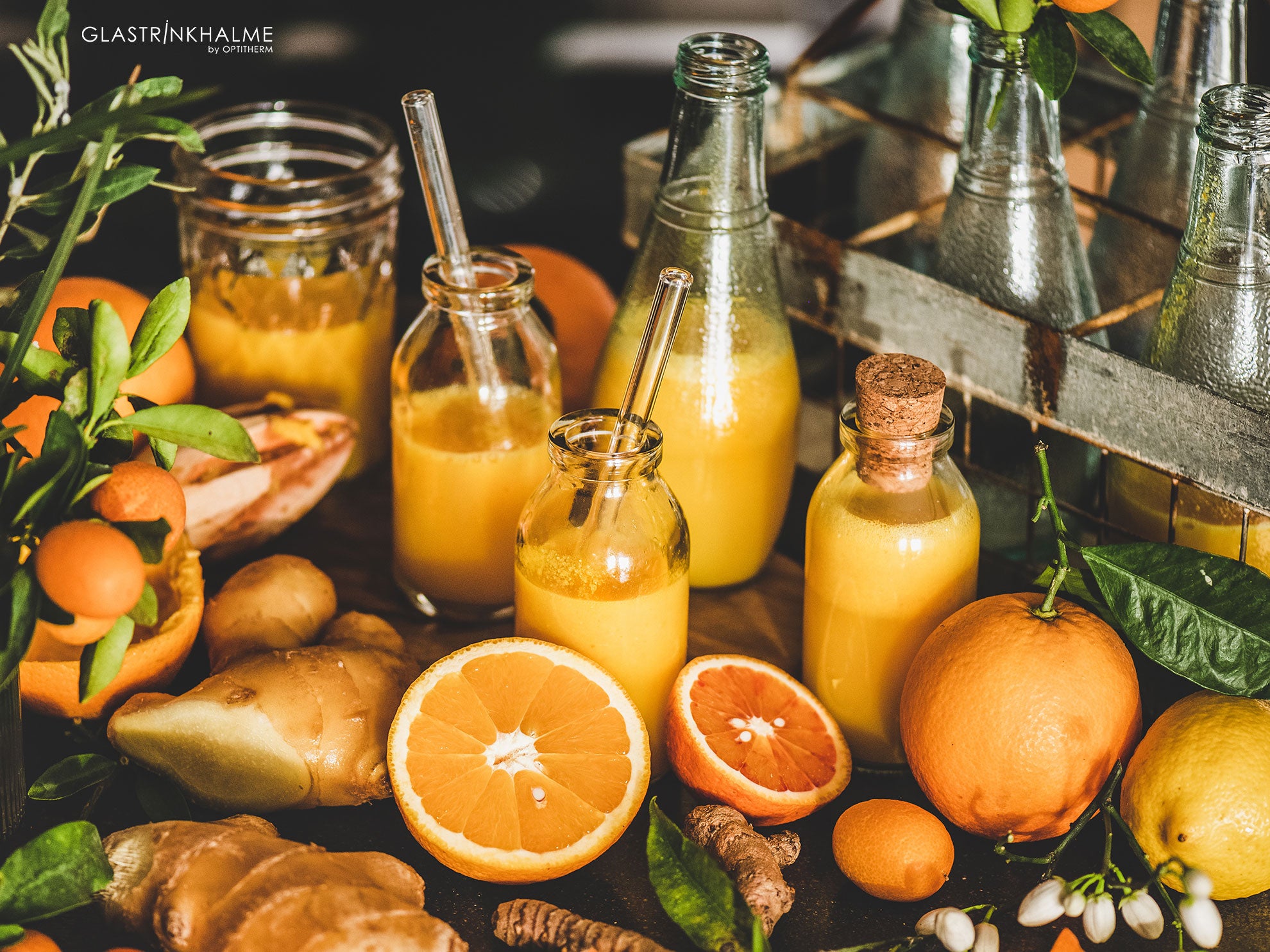 Ingwershots in einer kleinen Glasflasche mit Glastrinkhalm - rundherum liegen Orangen, Zitronen, Kurkuma, Gläser und gefüllte Glasflaschen