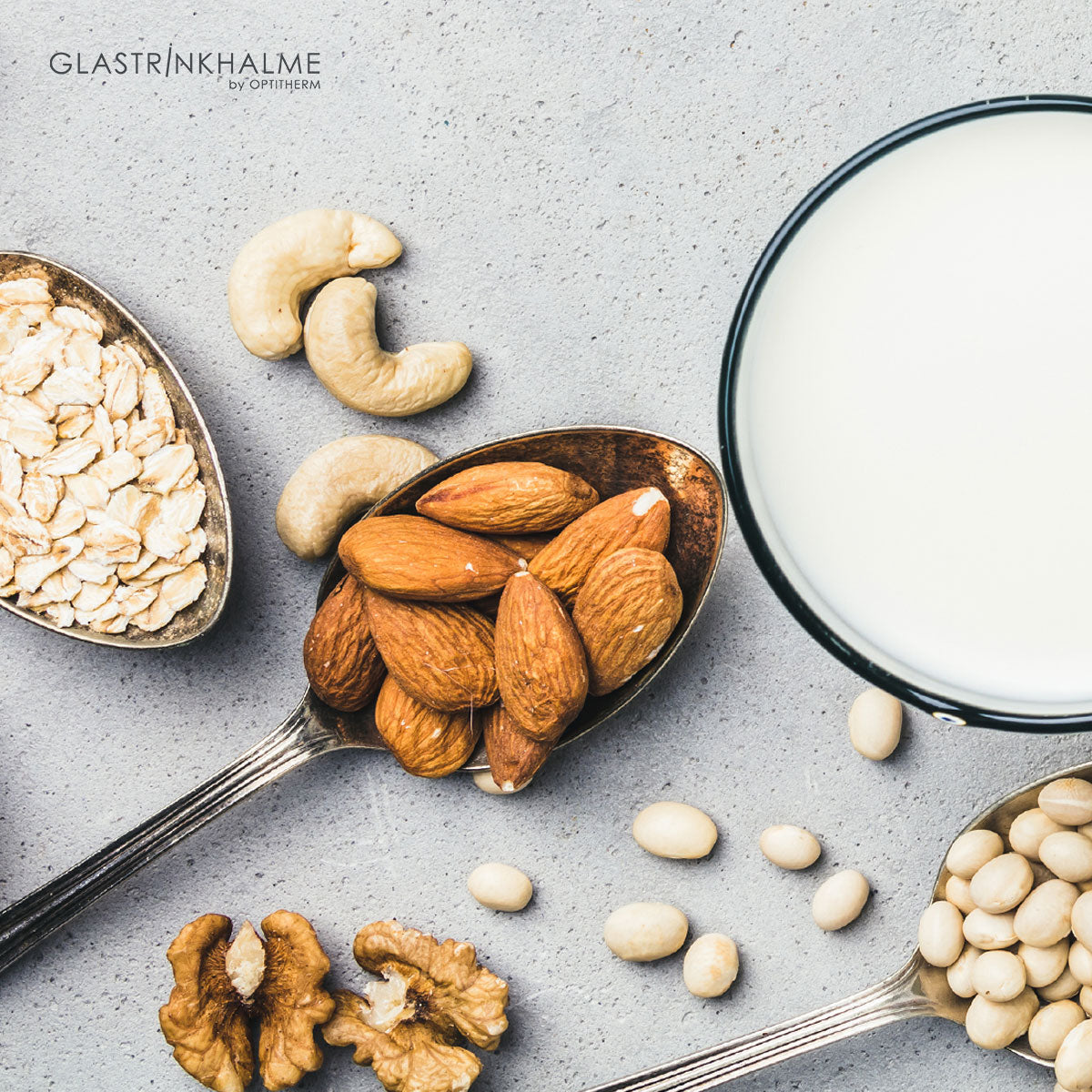 Ein Glas von oben mit alternative Milch mit Mandelkerne, Haferflocken, Linsen auf jeweils einem Löffel und verstreute Caschewnüsse, Walnusskerne und Linsen