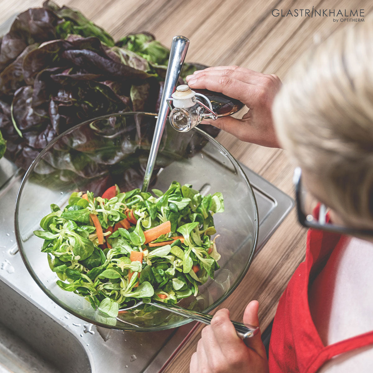 Frau mit roter Schürze bereitet einen grünen Salat in einer Glasschüssel zu