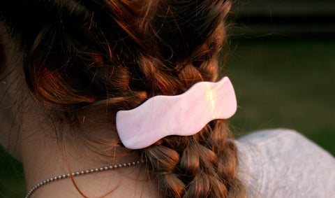 The back of a red haired person's head with their hair in braids and a pink iridized glass barrette clipped in at the nape of their neck.