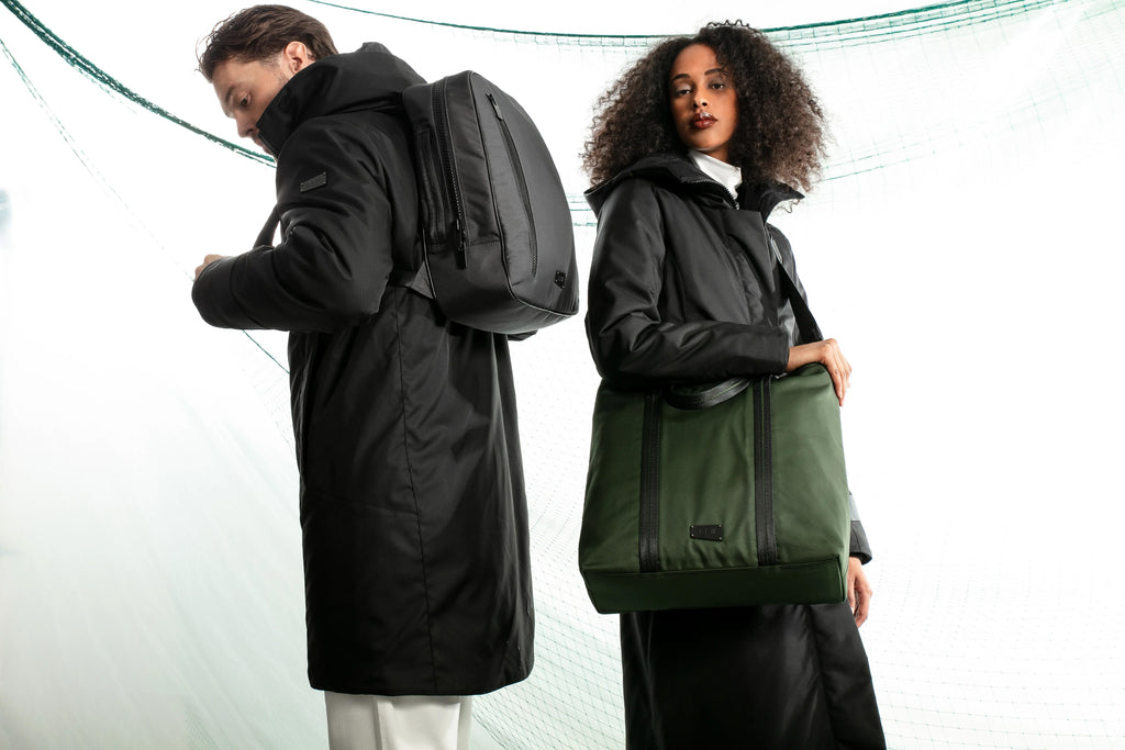 male model wearing a parka and carrying a backpack (left) a female model wearing an extended parka with a green tote