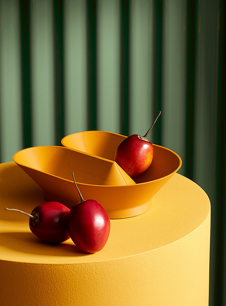 lifestyle picture of a yellow plastic fruit bowl with exotic red fruits near/inside it. the item is displayed on a yellow cylindrical table and in front of green show curtains