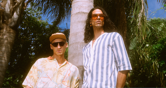 Two men wearing shirts and sunglasses standing in front of palm trees, exuding a summer vibe