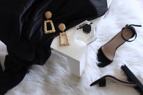 Statement gold squared hoop earrings laid across a black black dress and placed besides a perfume and black heels.
