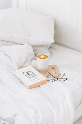 A cup of coffee, a book and a pair of sunglasses laid on a tray on top of a white duvet and pillow on a bed.